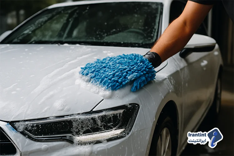 شست‌وشوی بدنه یک خودروی سفید با دستکش میکروفایبر آبی و کف فراوان در حال تمیزکاری